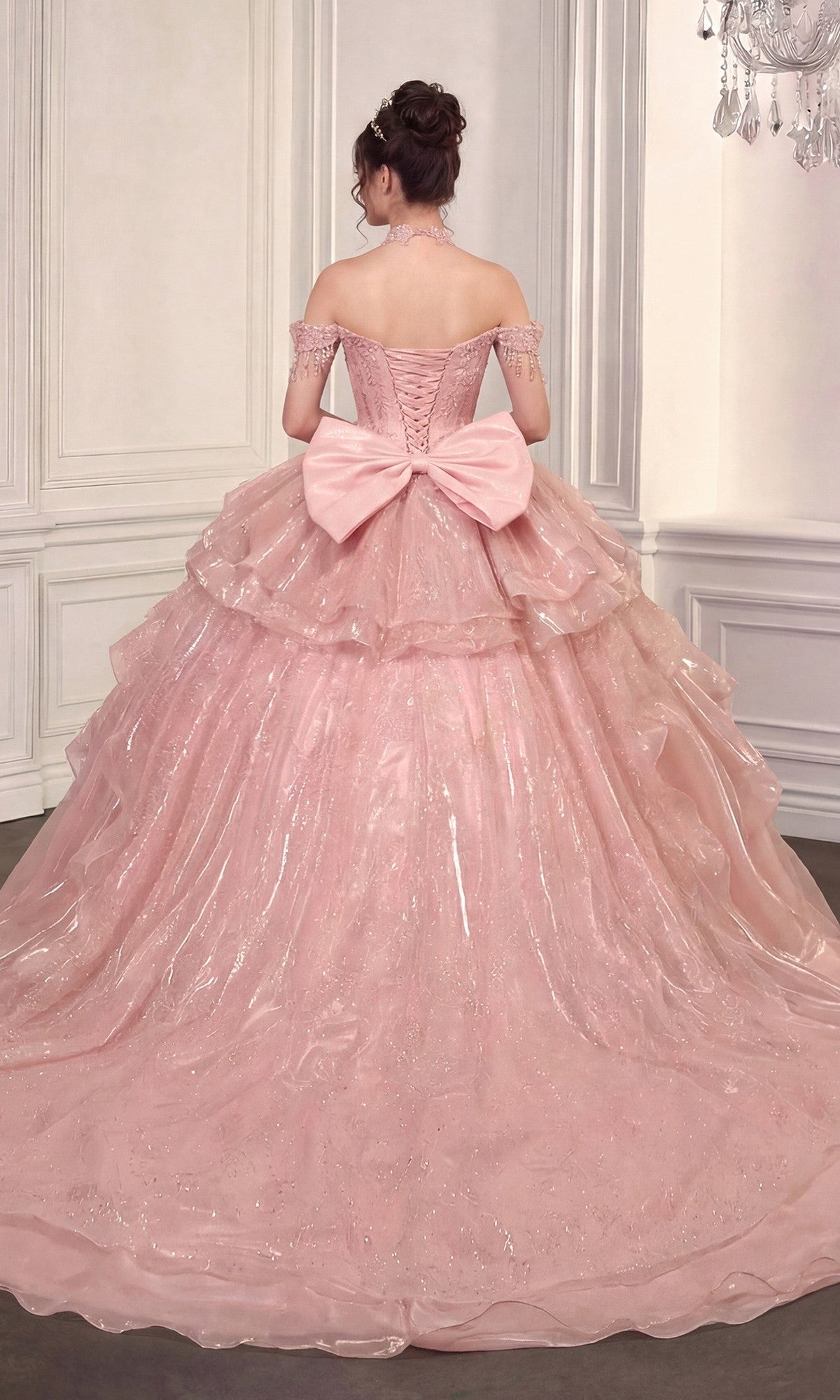 A woman wears the Quinceanera Dress 15742 By Ladivine—an off-the-shoulder pink gown with a detachable skirt and large back bow—while standing in an ornate room with white paneled walls, facing away.